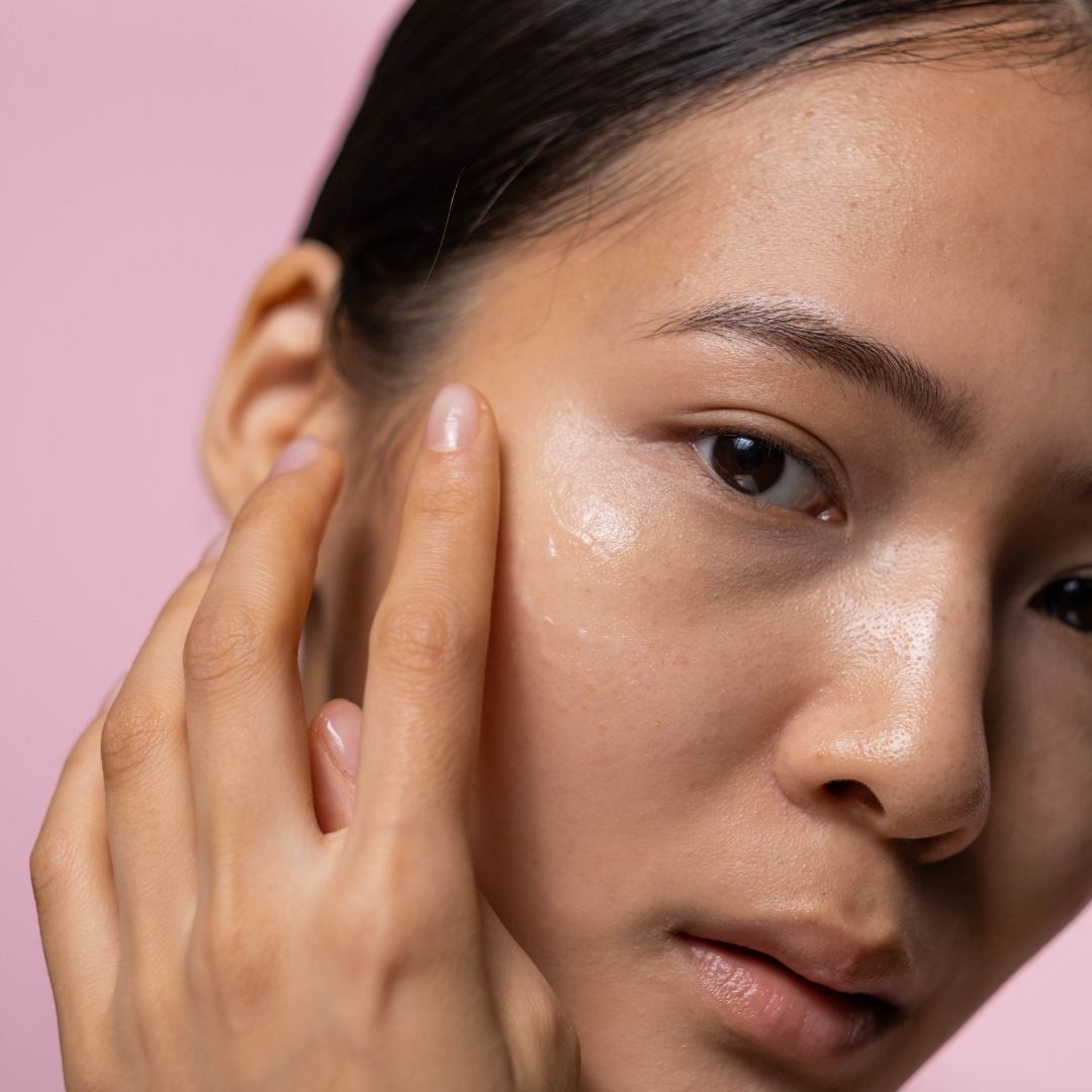 Close-up of a person applying skincare product to their face against a pink background